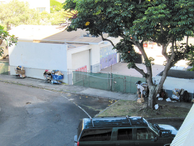 A homeless man (under tree) examines his belongings, which extend the length of a Kailua sidewalk in a metered city parking lot next to the now-closed Kuulei Union service station. Photo by Claudia Webster. 
